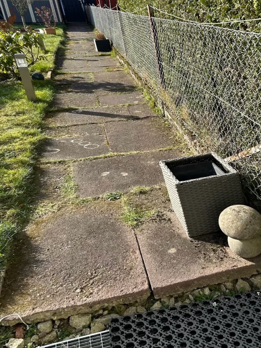 Vorher: Weg aus verwitterten, bemoosten Betonplatten neben einem alten Maschendrahtzaun